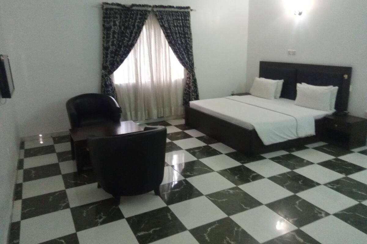 A hotel room with a checkered floor, a bed with white linens, a desk with a chair, and a window with patterned curtains.