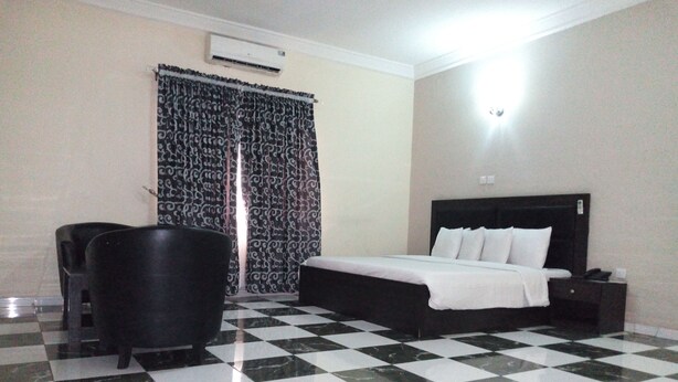 A hotel room with a checkered floor, a bed with white bedding, a black headboard, a nightstand, a black chair, and patterned curtains. - Eliata Hotel & Suites (Lagos)