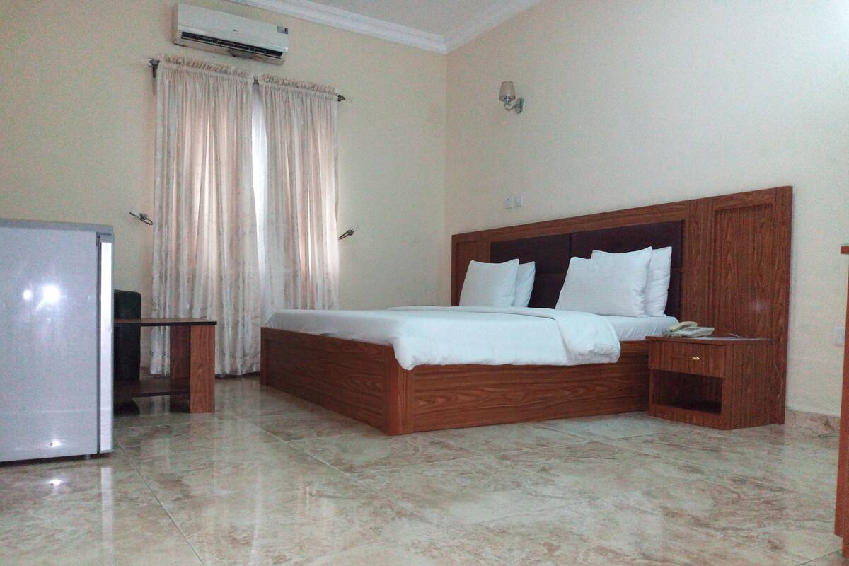 A bedroom with a wooden bed, a nightstand, a desk, a chair, a refrigerator, and a window with curtains.