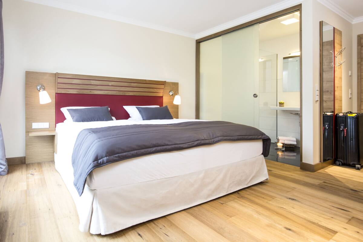 Quarto de hotel moderno com cabeceira de madeira, uma cama grande com cobertura cinza e branca, mesa de cabeceira com abajur e porta de vidro deslizante para um banheiro.