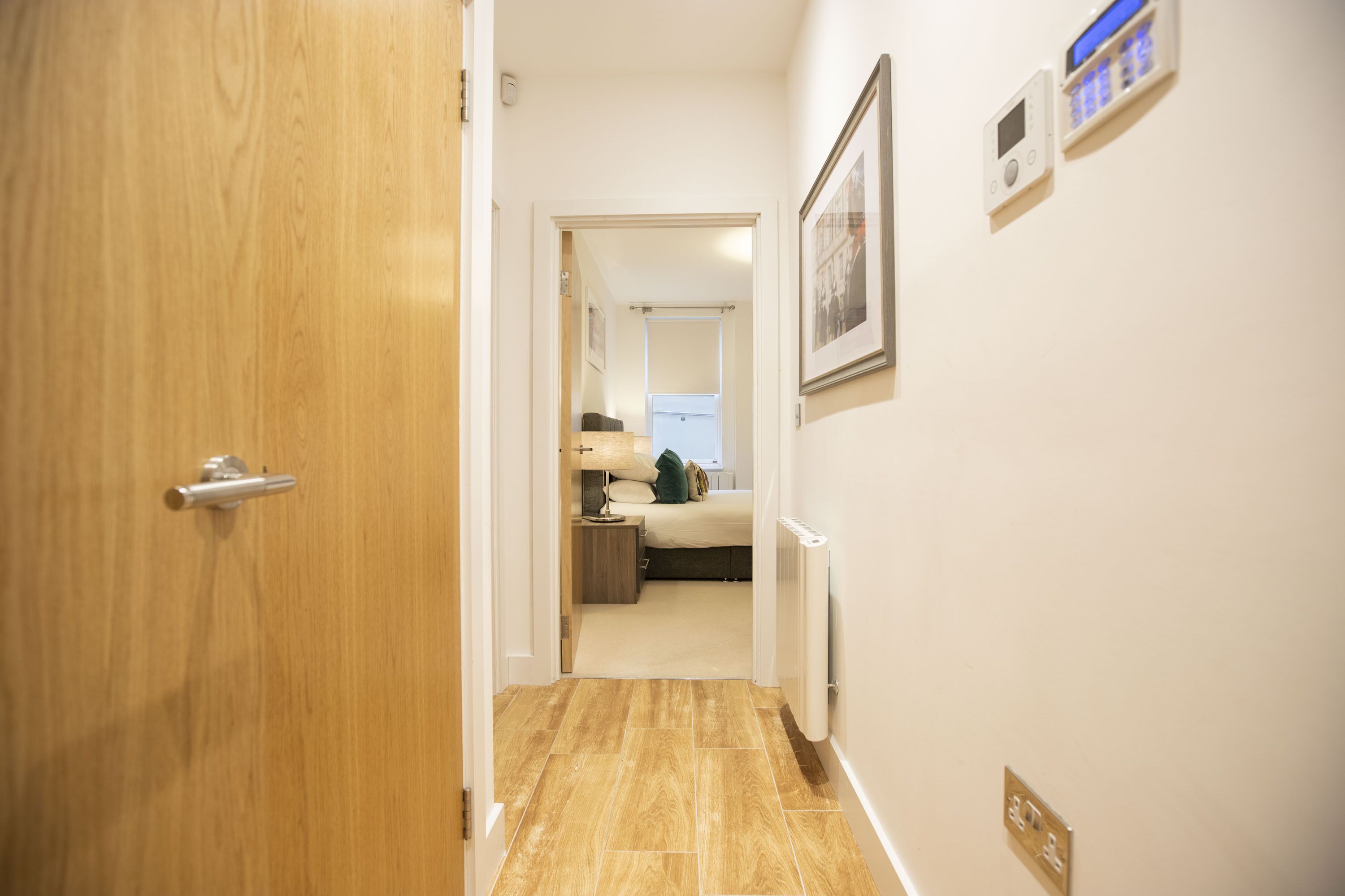 A hallway with a wooden door, a bed with a nightstand, a radiator, and a framed picture on the wall.