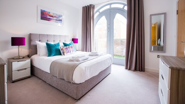 A modern bedroom with a large bed, two bedside tables, a nightstand, a dresser, a mirror, and a window with curtains. - King Edward Apartments (Windsor)