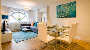 A modern living room with a glass dining table, white chairs, a sofa with colorful cushions, a large window with curtains, and a painting on the wall. - King Edward Apartments (Windsor)