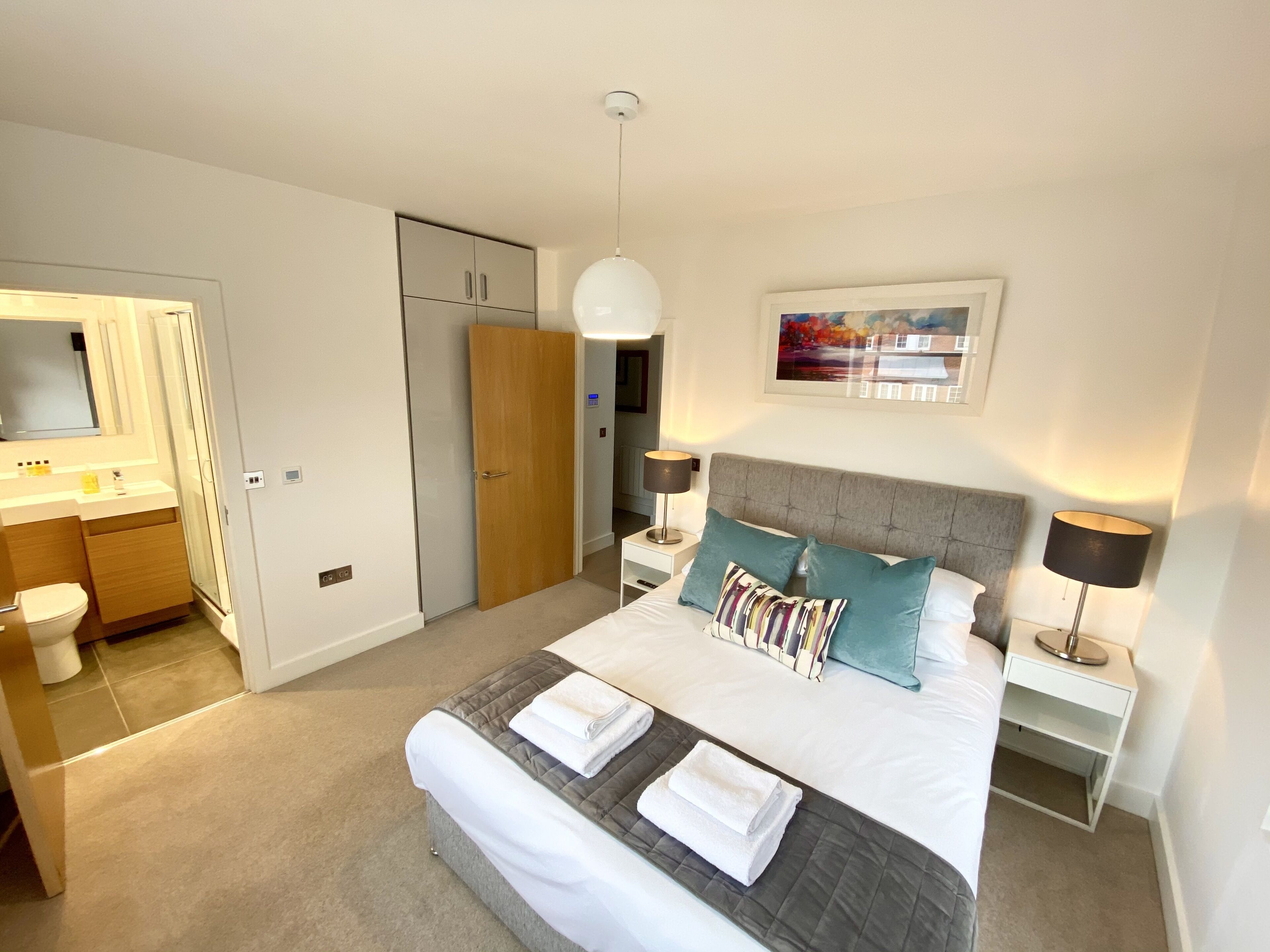 A modern bedroom with a large bed, bedside tables, a nightstand, a mirror, a wardrobe, and a bathroom.