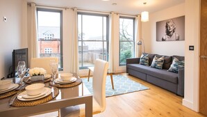 A modern living room with a grey sofa, a dining table set for four, and large windows offering a view of buildings and trees. - King Edward Apartments (Windsor)