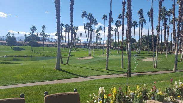 A golf course with palm trees, a pond, and a building in the background. - Palm Valley with Beautiful View and Platinum Membership (Palm Desert)