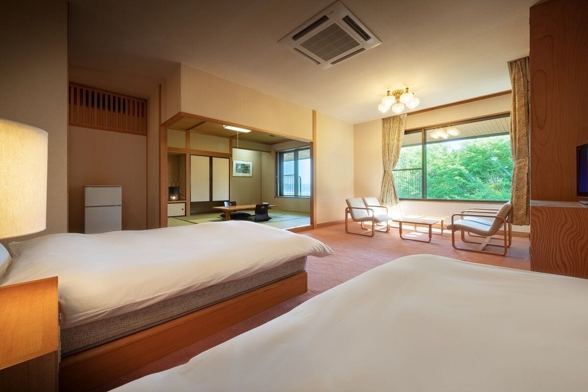 Une chambre d’hôtel avec deux lits, un bureau, des chaises, une télévision et une grande fenêtre donnant sur un paysage verdoyant.
