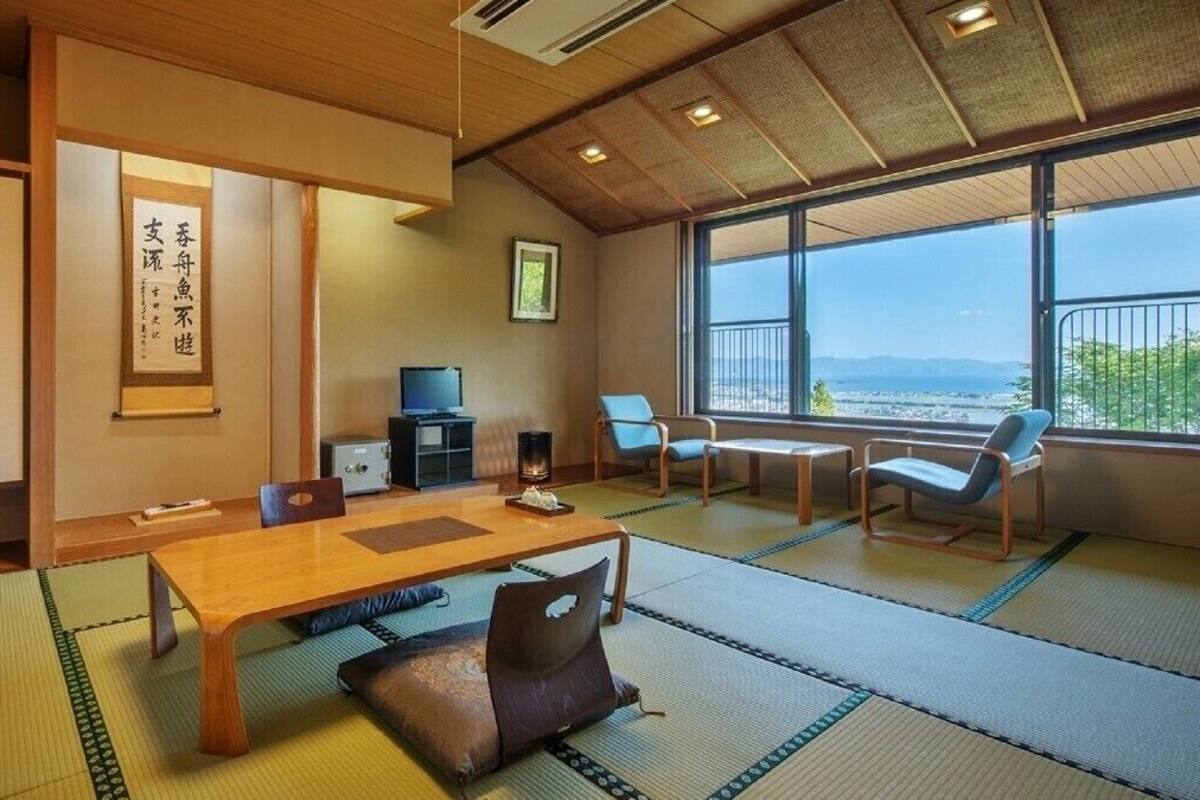 Une pièce de style japonais traditionnel, avec un sol recouvert de tatamis, une table en bois, des chaises, un téléviseur et une vue dégagée sur l’extérieur grâce à de grandes fenêtres.