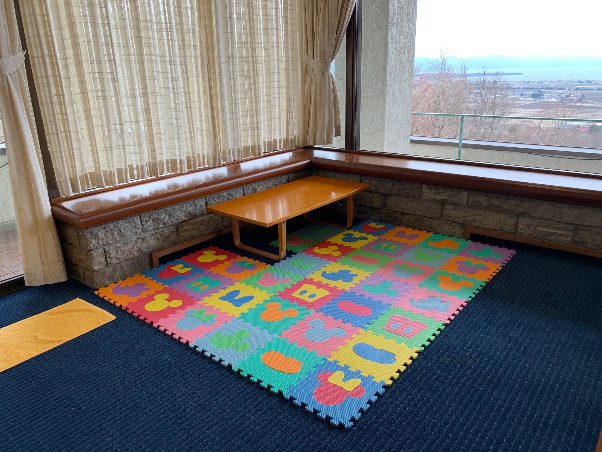A corner room with a bench, a colorful play mat, and a view of a distant landscape through a window.