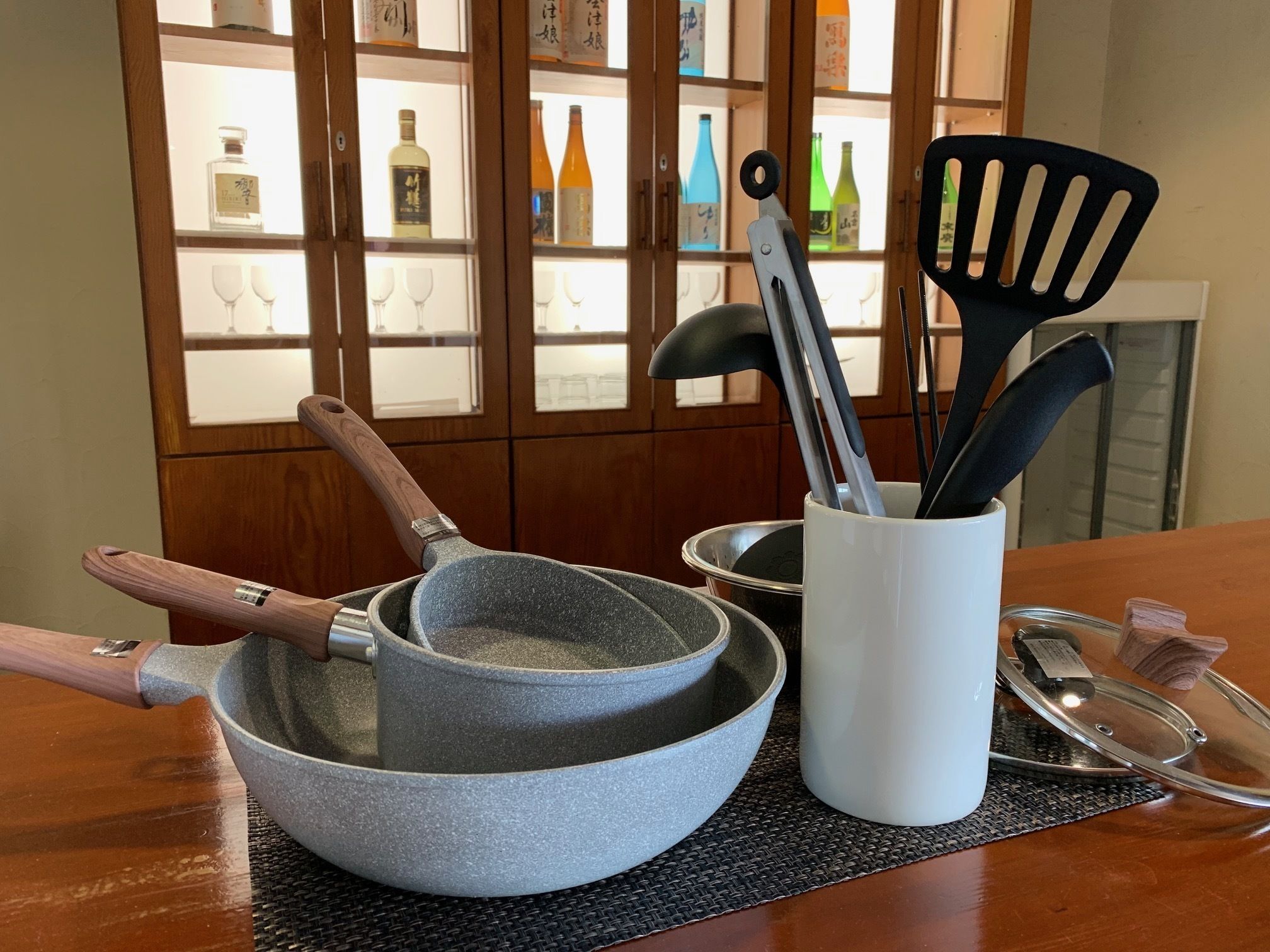 A kitchen counter with a stone mortar and pestle, a wooden-handled pan, a white mug with utensils, and a cabinet with bottles in the background.
