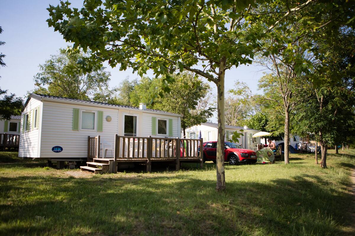 A mobile home with a deck, a red car parked nearby, and a tree in the foreground.