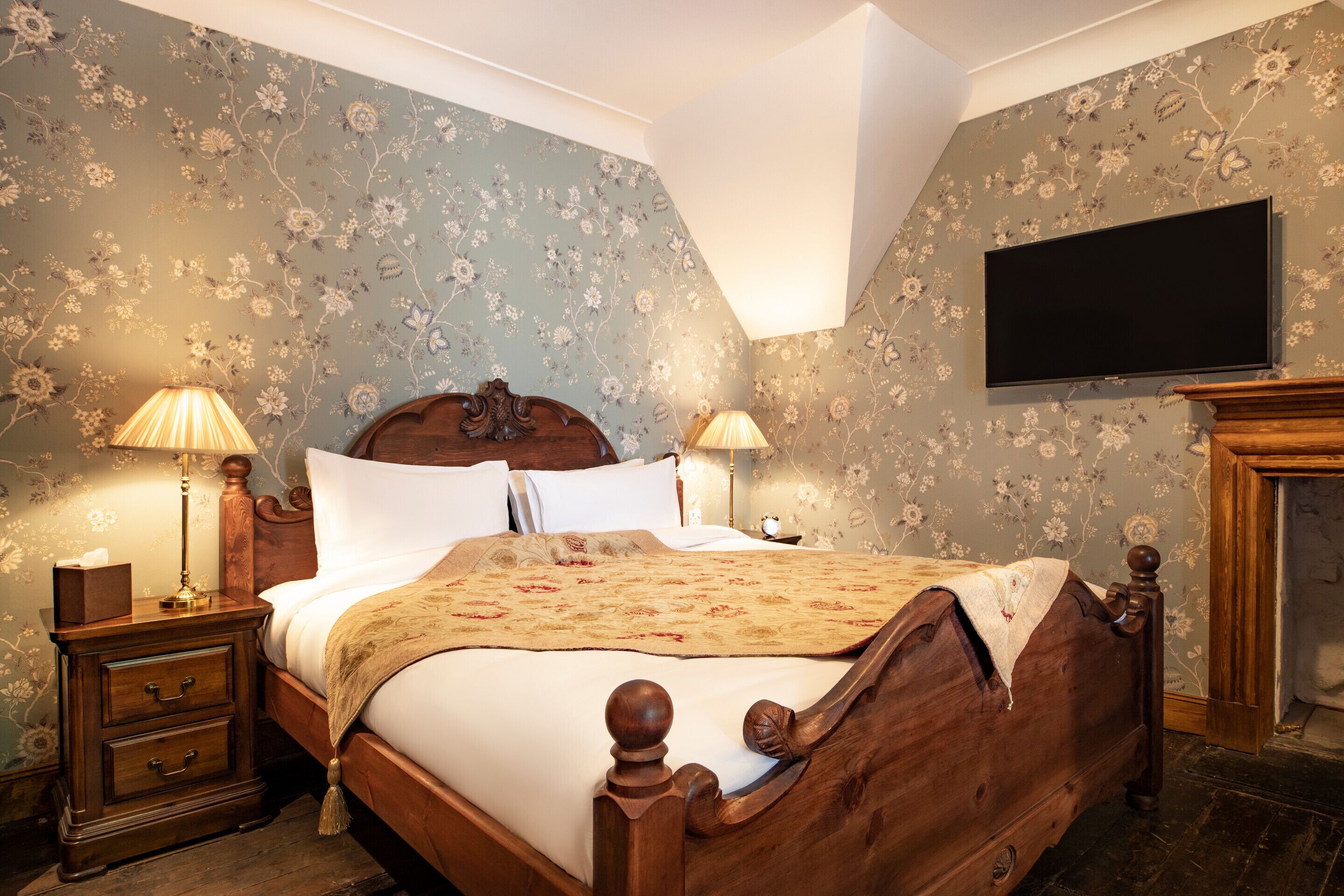 Un dormitorio con cama de madera, dos mesitas de noche con lámparas, un televisor de pantalla plana montado en la pared y papel tapiz con motivos florales.
