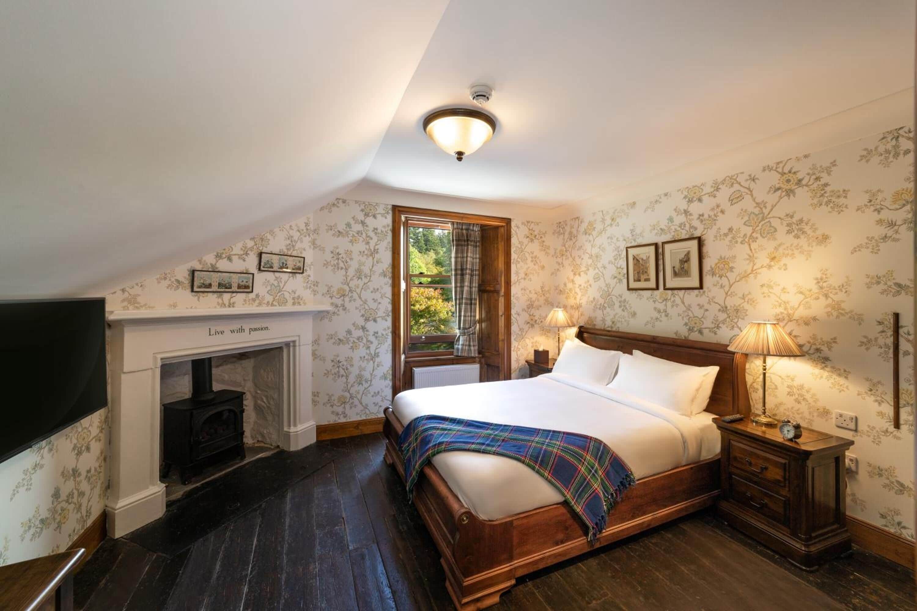 A bedroom with a wooden bed, a fireplace, a nightstand, and a window with a view of greenery.