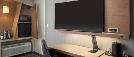 Habitación de hotel con televisión de pantalla plana montada en la pared, escritorio con silla, microondas, refrigerador pequeño y calefacción.