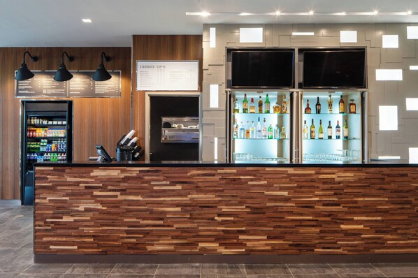 A modern bar area with a wooden reception desk, a menu board, a beverage dispenser, and a refrigerator stocked with drinks. - Courtyard by Marriott Lafayette South (Lafayette)