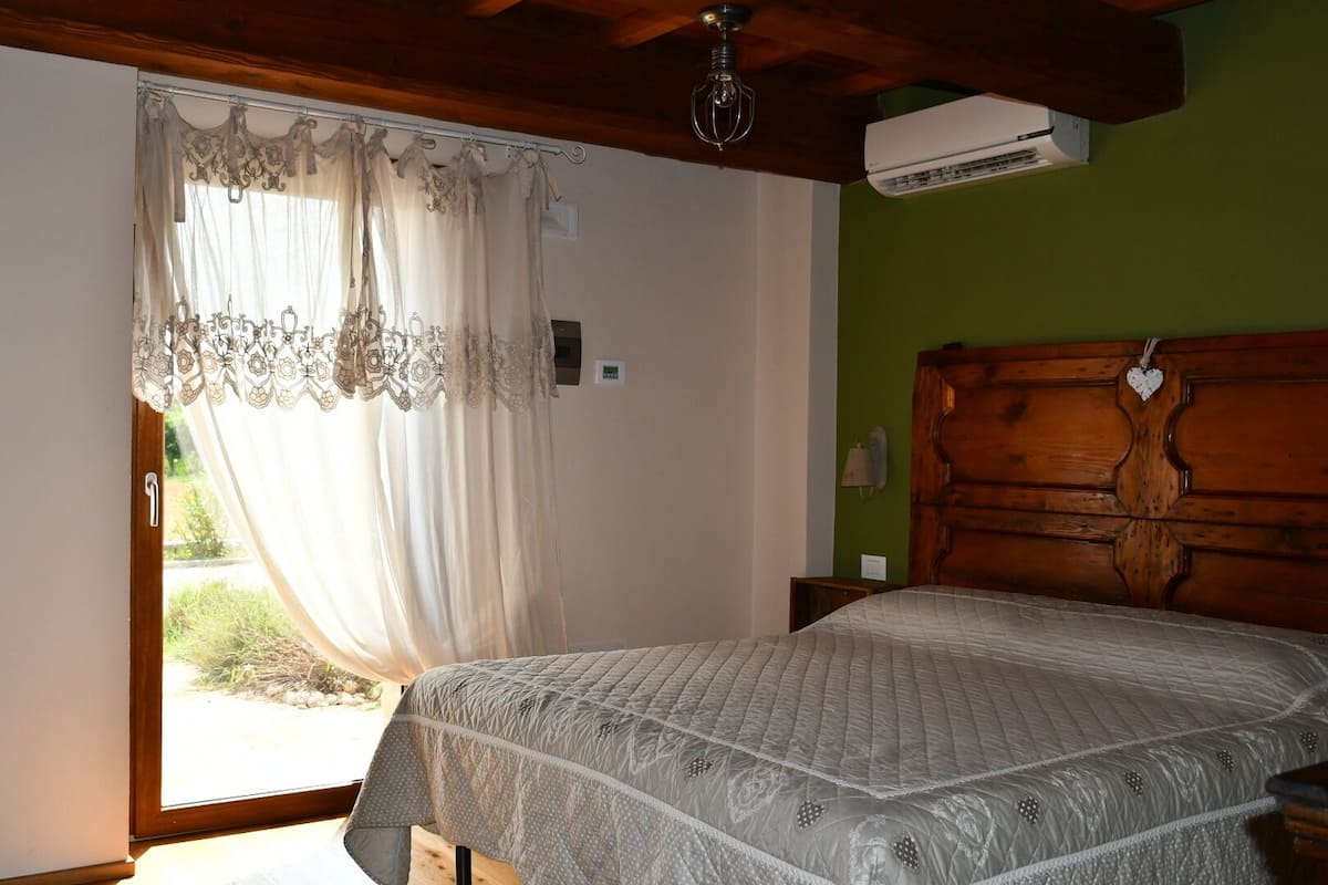 Une chambre à coucher comprenant un lit, une tête de lit en bois, un appareil de climatisation, une fenêtre avec des rideaux et une petite table de chevet.