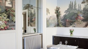 Un comedor con mesas y sillas de mármol, un espejo grande y un mural con un paisaje.