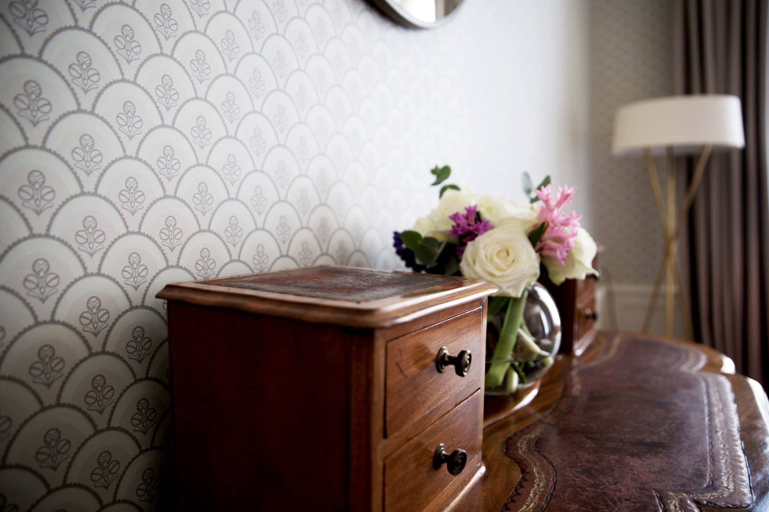 A wooden dresser with two drawers, a vase with flowers, and a patterned wallpaper.