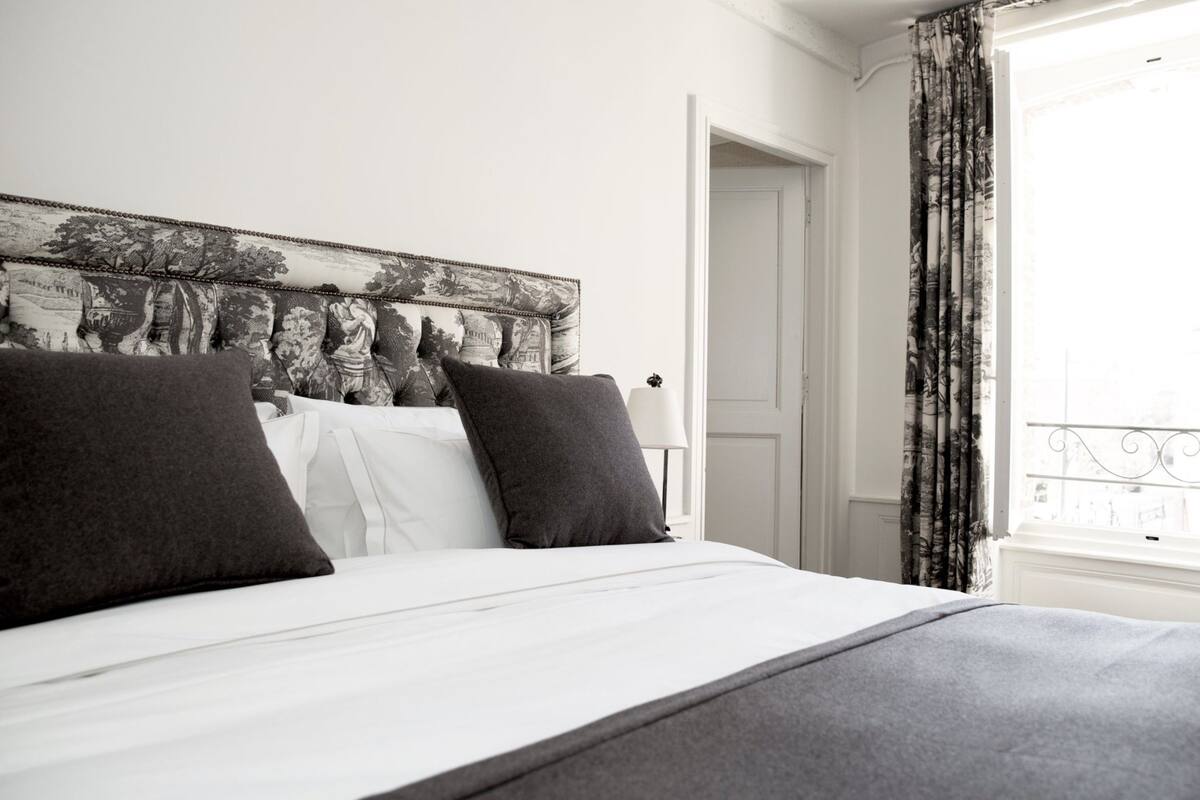 Una cama bien hecha con cabecera blanca y dos almohadas oscuras, una puerta blanca y una ventana con vistas a un balcón.