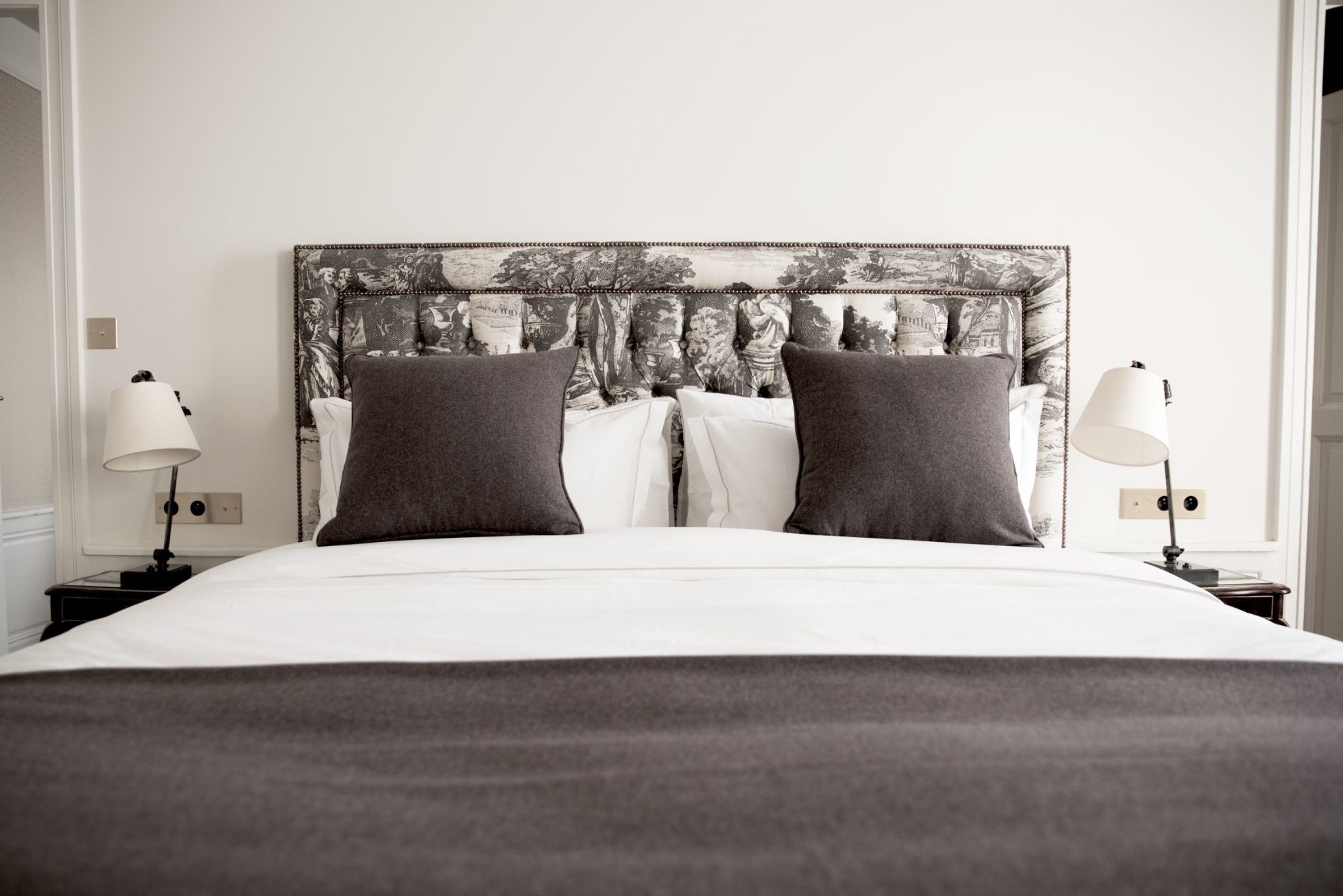 A neatly made bed with a large headboard, two bedside lamps, and a wall with a patterned tapestry.