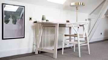 Una habitación blanca con techo inclinado, un radiador, un cuadro enmarcado, una mesita, una silla y una lámpara de pie.