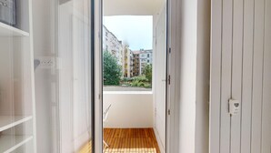 Porta aperta che dà su un balcone con pavimento in legno, affacciato su una zona residenziale con edifici e vegetazione.