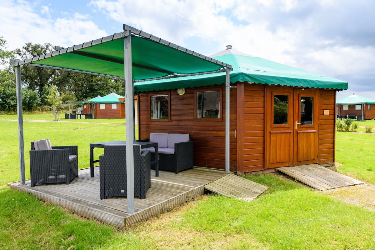 A wooden cabin with a green roof, a porch with wicker furniture, and a ramp leading to the entrance.