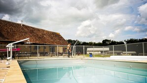 Una piscina con solárium de baldosas, cercada por una valla metálica, y un edificio con tejado de cerámica al fondo.