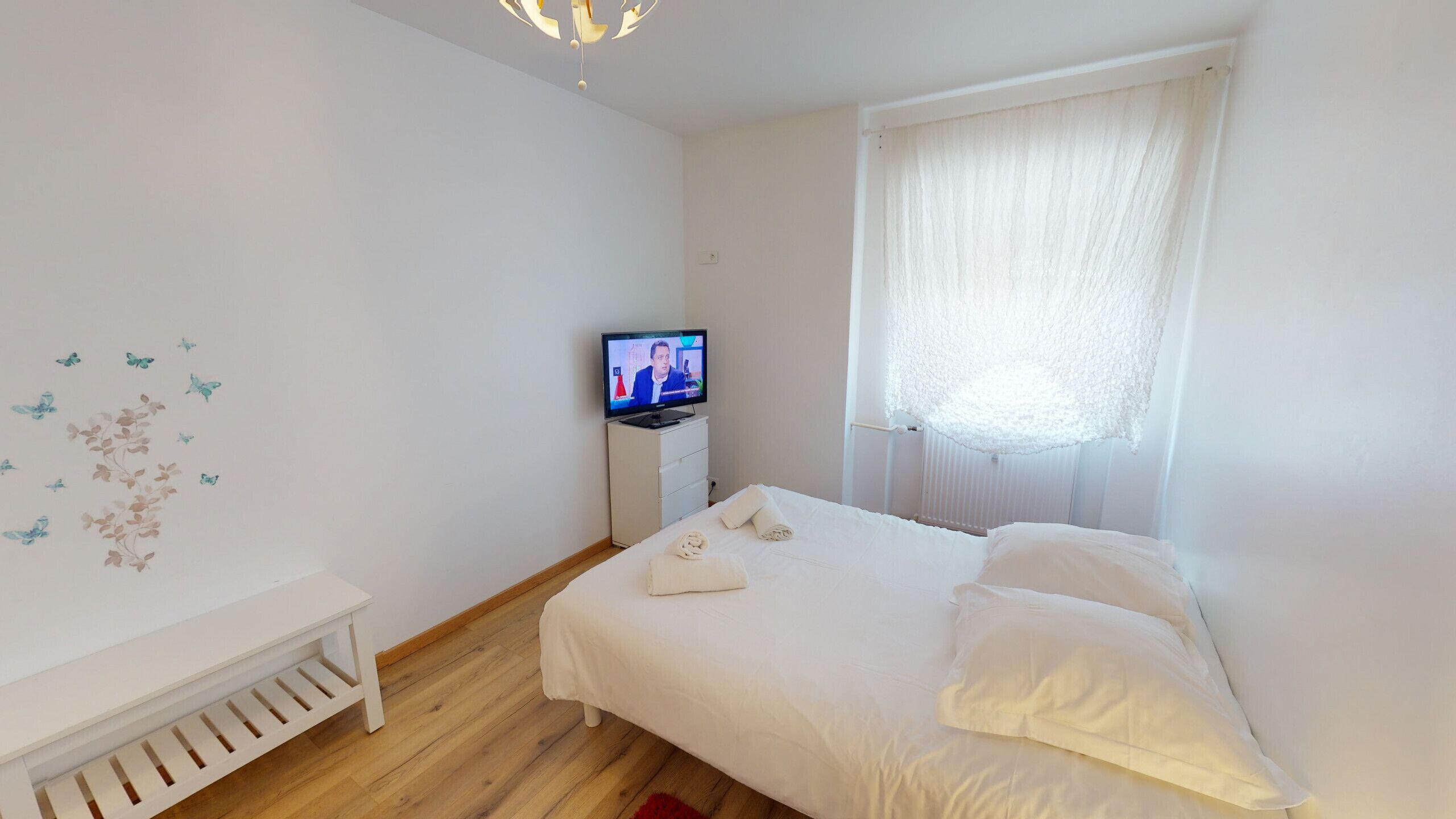 Een slaapkamer met een bed, een televisie, een witte ladekast en een raam met gordijnen.