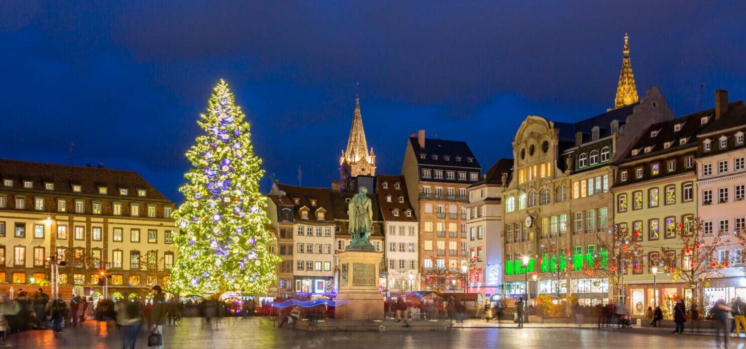 Une place historique, ornée d’un grand sapin de Noël et d’une statue, et entourée d’édifices représentatifs de l’architecture européenne traditionnelle.