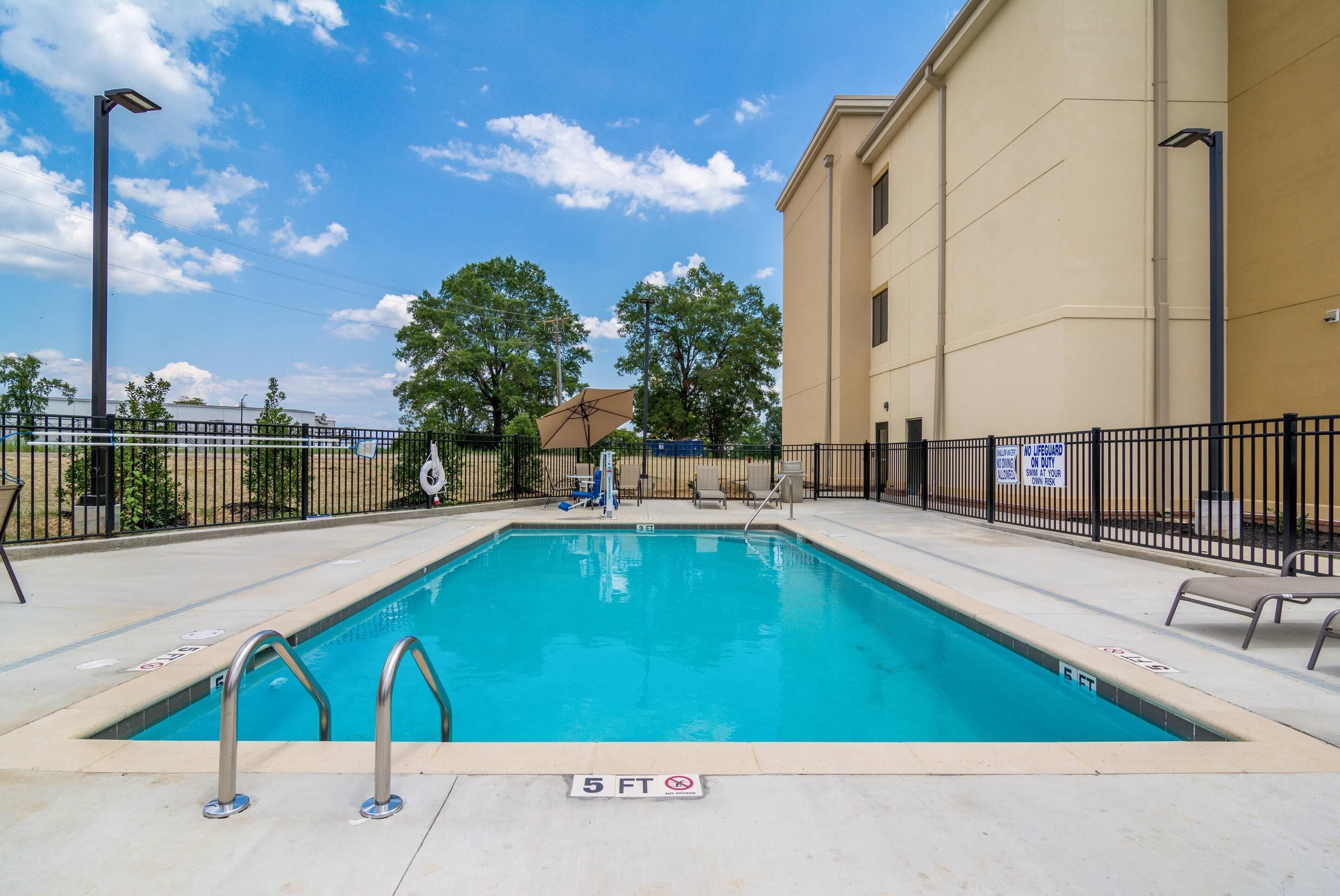 A swimming pool with a 5 ft depth marker, surrounded by a fence, with a building and trees in the background.