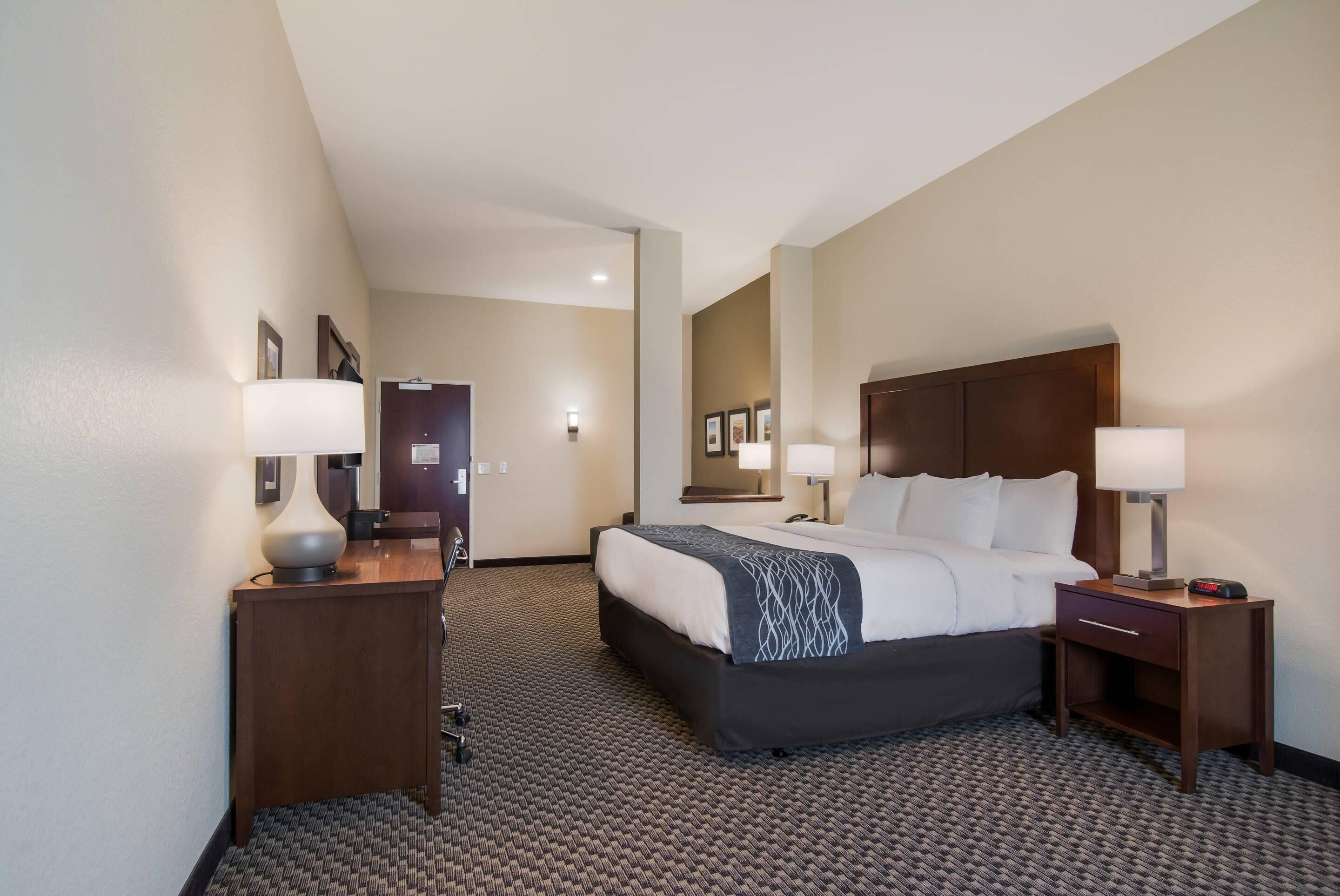 A hotel room with a bed, two bedside tables, a nightstand, a lamp, a mirror, and a carpeted floor.