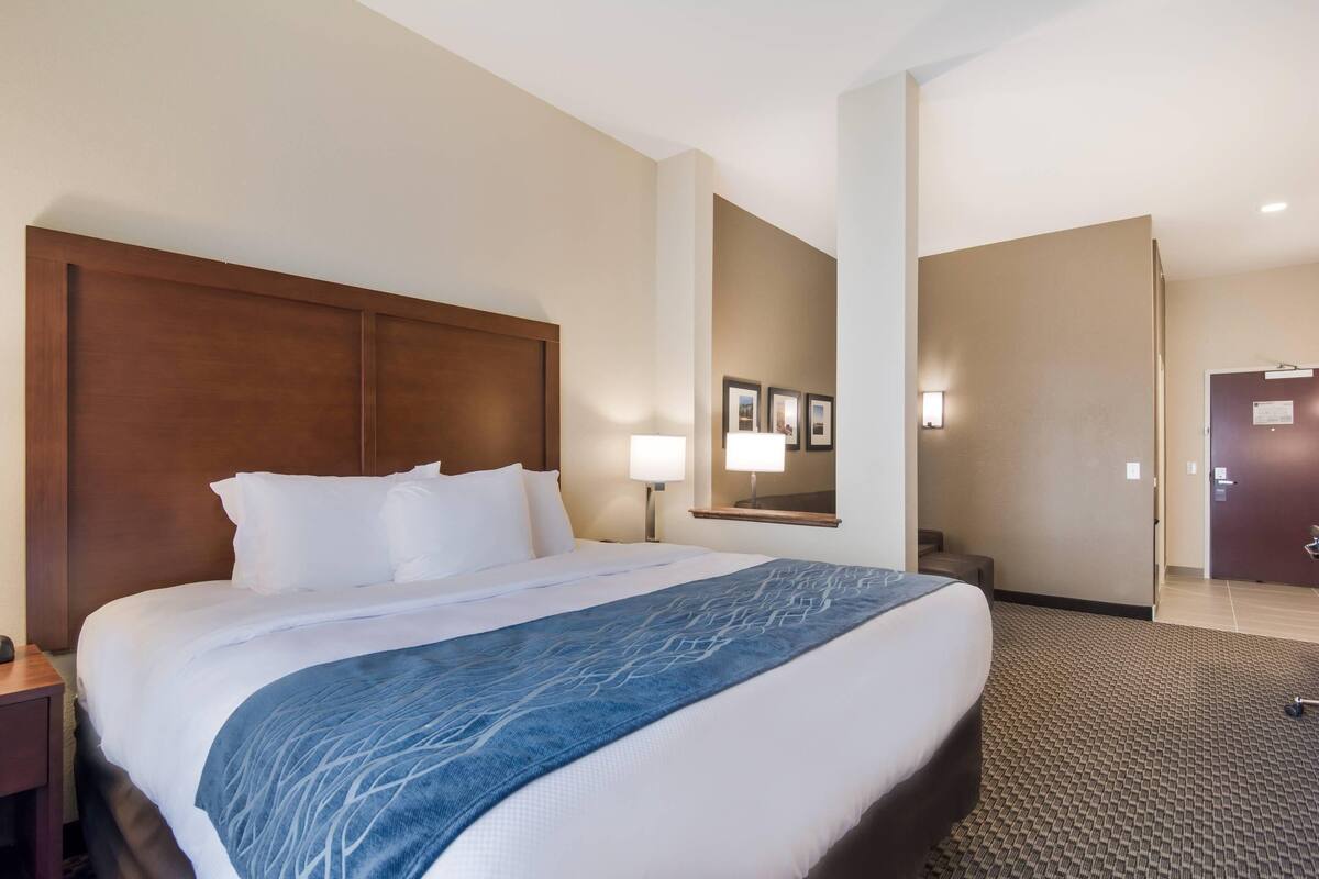 Ein Hotelzimmer mit einem großen Bett, einem Holz-Kopfteil, einem Nachttisch, einer Lampe, einem Spiegel und einem Teppichboden.