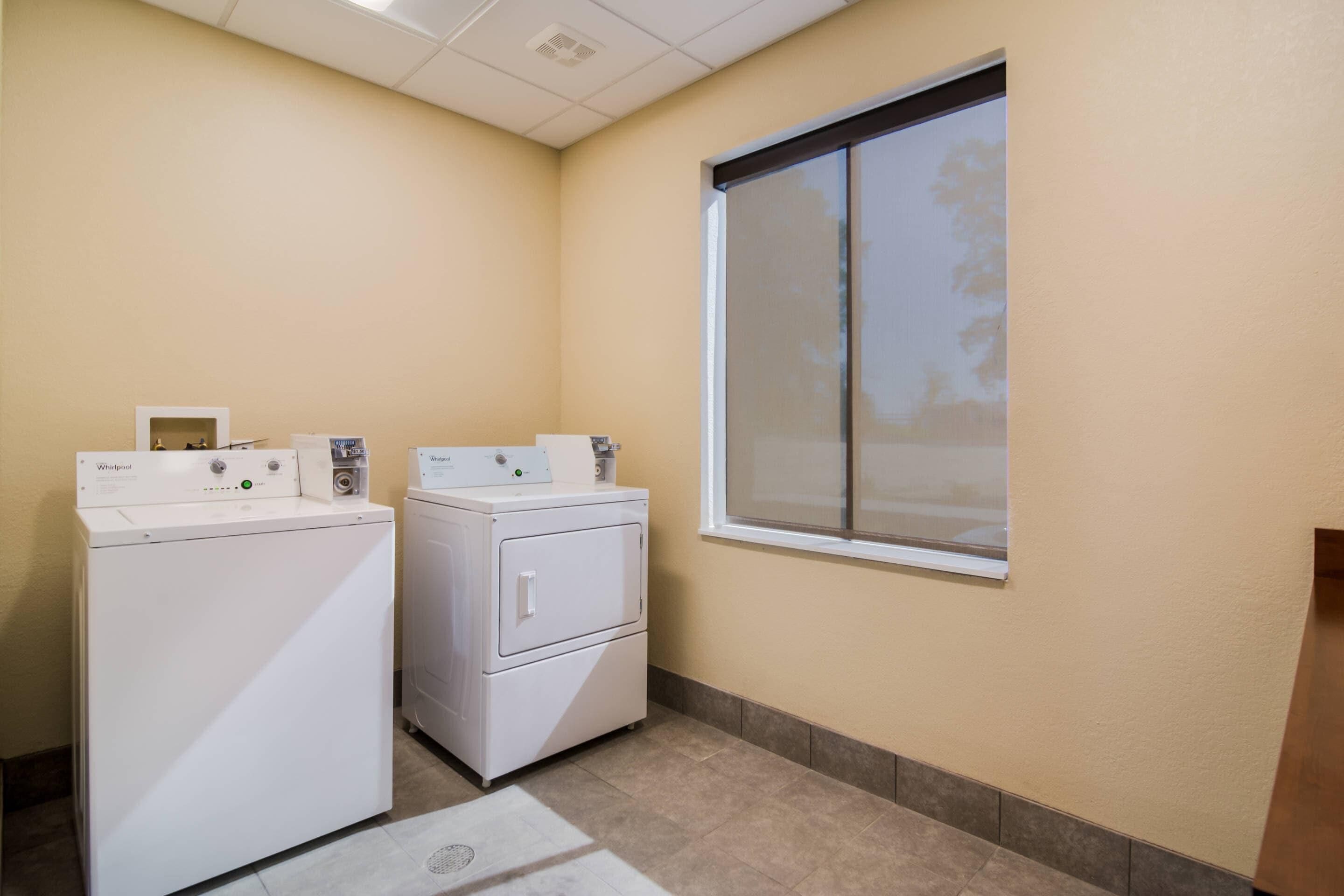 A laundry room with two white Whirlpool washers and dryers, a window with blinds, and a tiled floor.