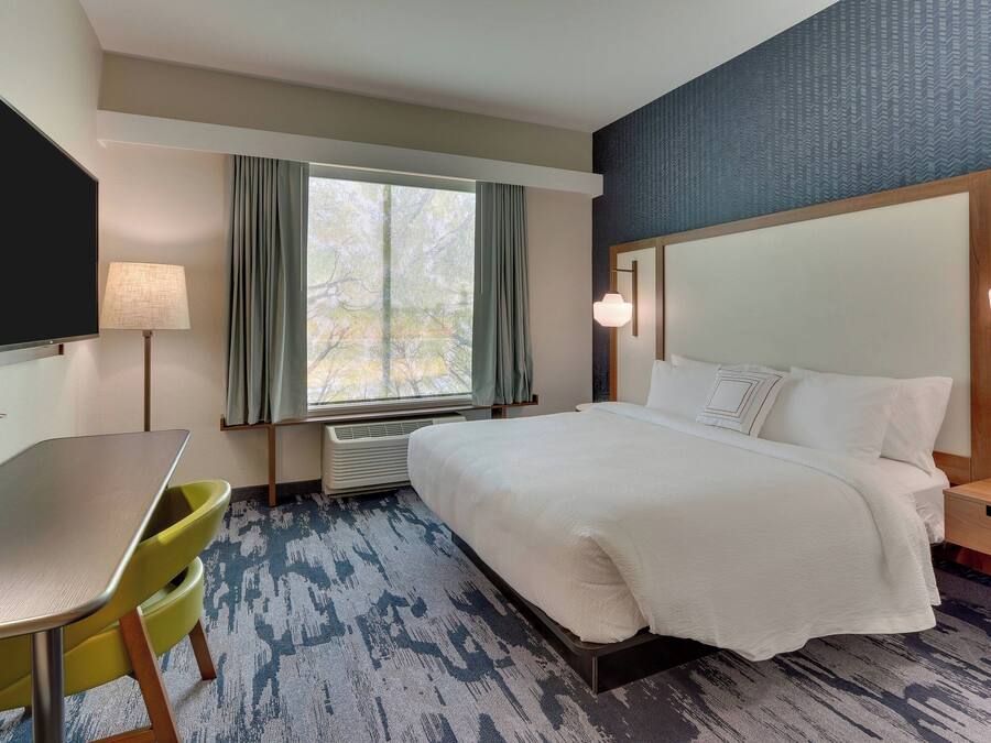 Habitación de hotel con cama, escritorio, silla, televisor y ventana con cortinas.
