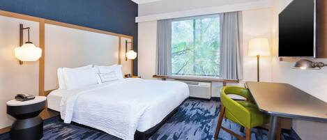 A hotel room with a large bed, a desk, a chair, a TV, and a window with a view of trees.