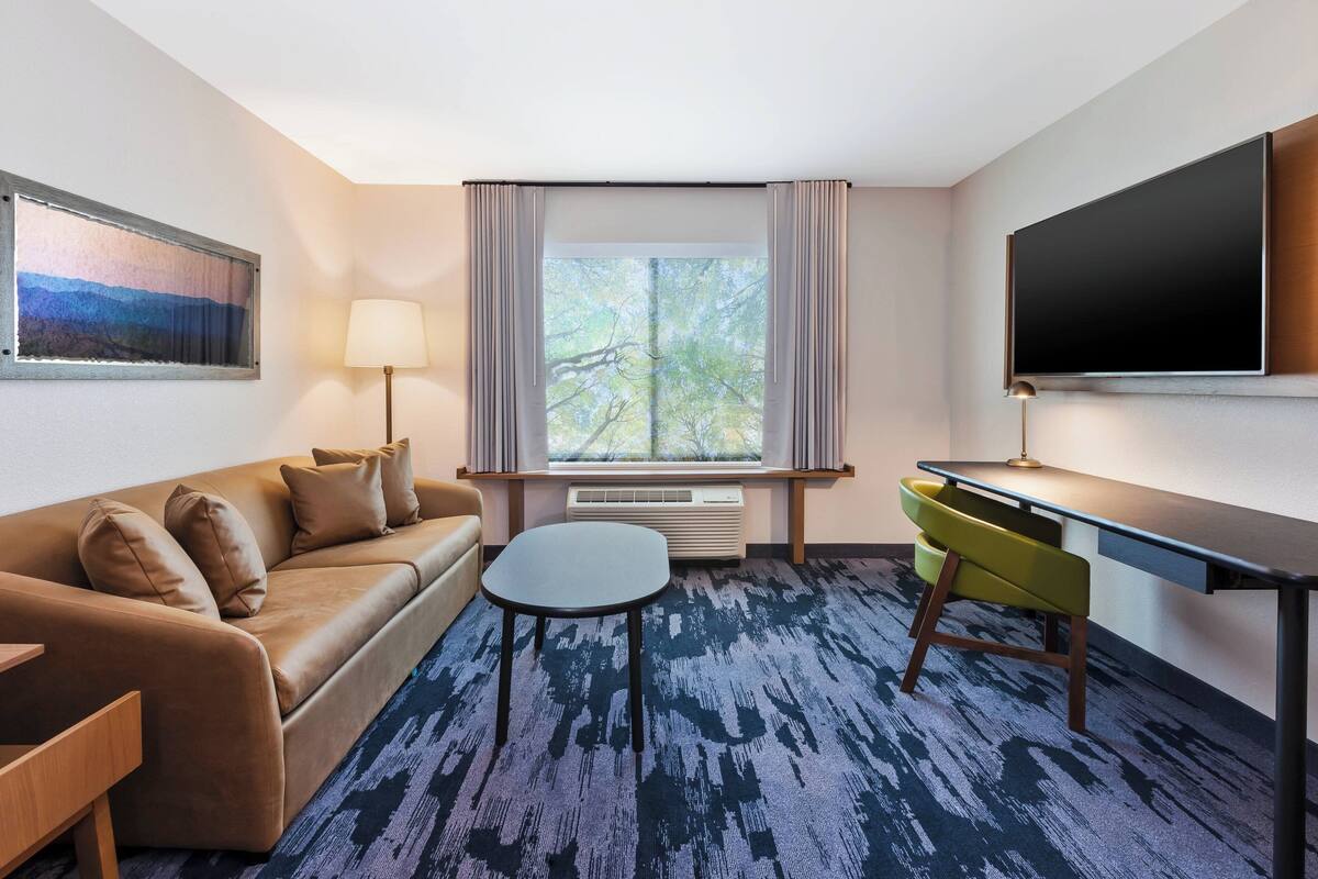 A hotel room with a sofa, a small table, a desk, a TV, a window with curtains, and a painting on the wall.