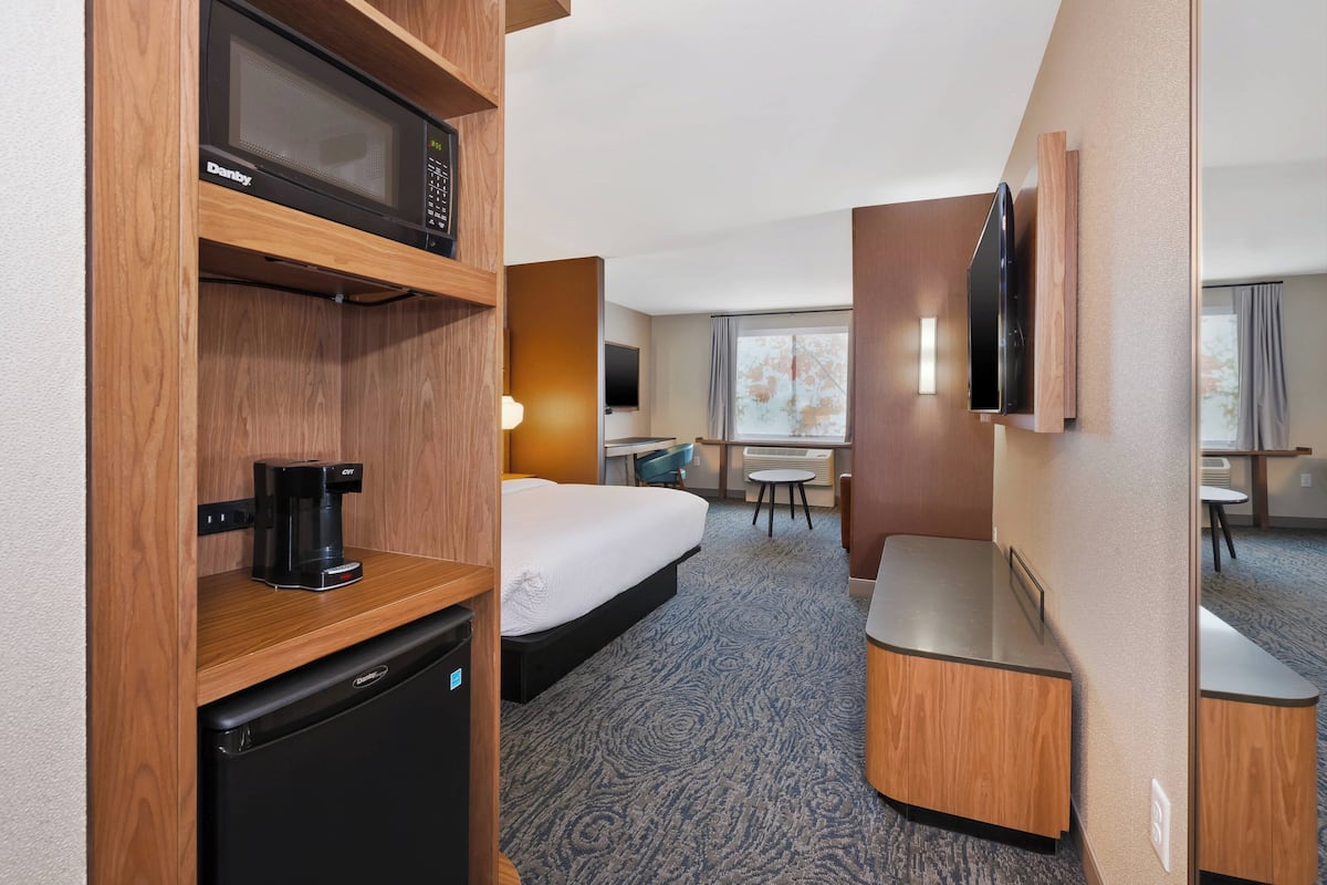 Ein Hotelzimmer mit Bett, Schreibtisch, Mikrowelle, Kaffeemaschine und einem an der Wand befestigten Fernseher.