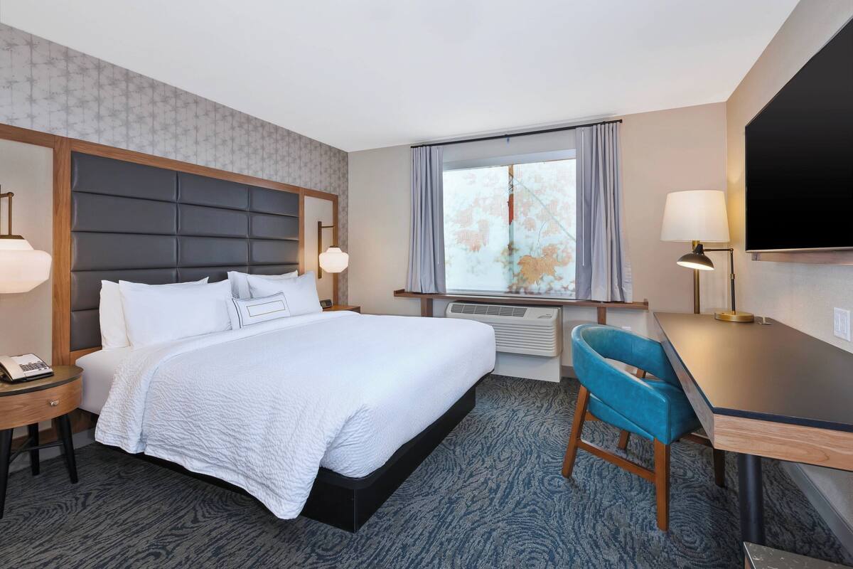 Ein Hotelzimmer mit einem großen Bett, einem Schreibtisch, einem Sessel, einem Fernseher und einem Fenster mit Vorhängen.