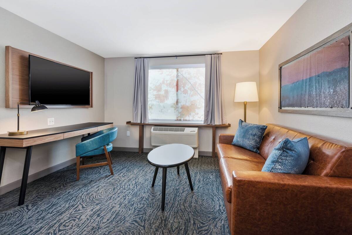 Ein Hotelzimmer mit Flachbildfernseher an der Wand, einem Schreibtisch mit Stuhl, einem kleinen runden Tisch, einem Ledersofa mit blauen Kissen, einer Stehlampe und einem großen Fenster mit Vorhängen.