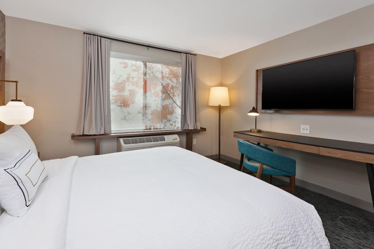 Ein Hotelzimmer mit einem großen Bett, einem Schreibtisch, einem Stuhl, einem Flachbildfernseher und einem Fenster mit Vorhängen.