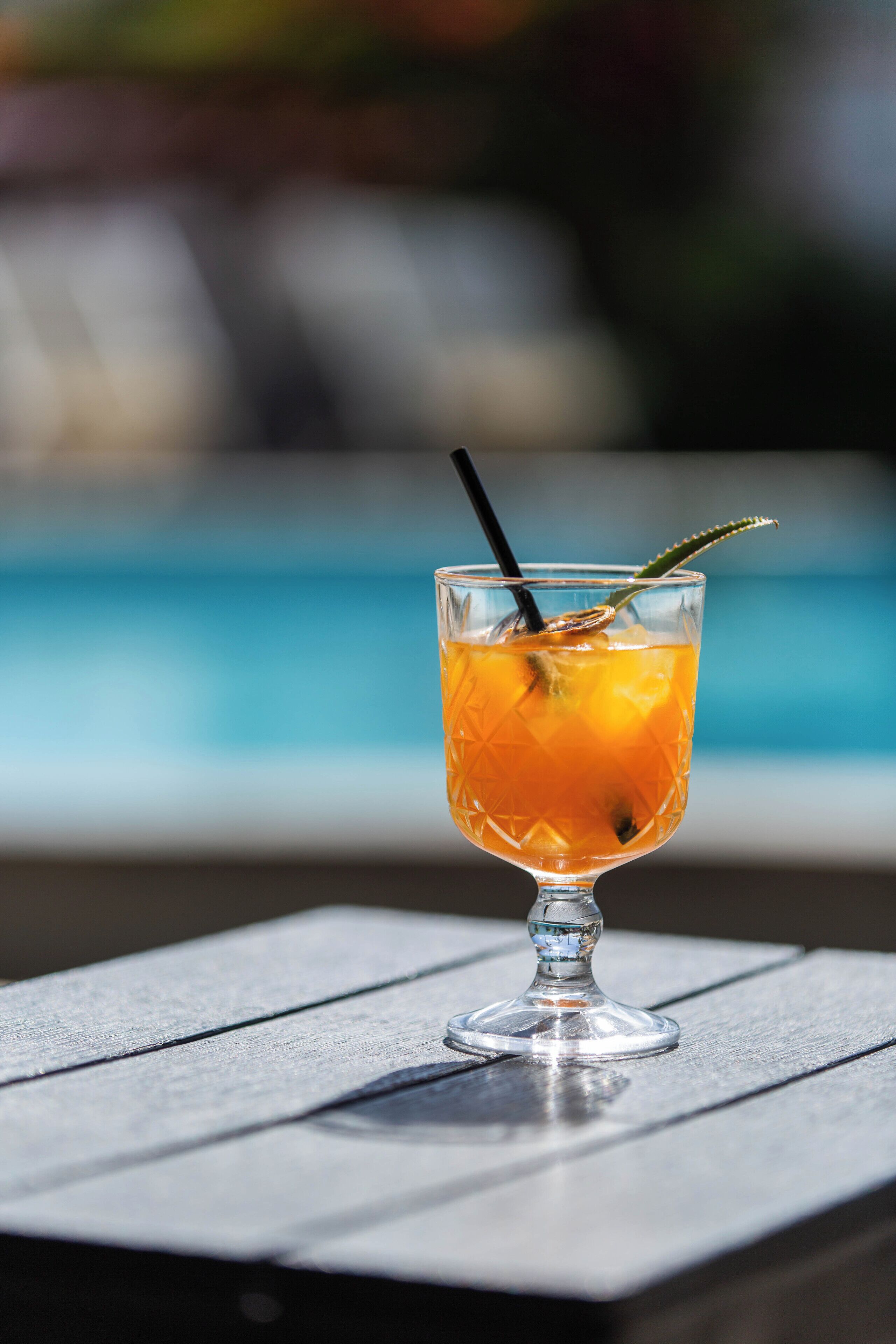 Un bicchiere da cocktail con una cannuccia e una guarnizione su un tavolo di legno vicino a una piscina.
