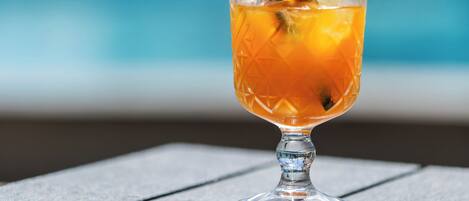 Un verre à cocktail avec une paille et une garniture, posé sur une table en bois près d’une piscine.
