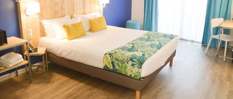 A hotel room with a wooden headboard, a bed with white and yellow bedding, a desk with a chair, and a window with blue curtains.