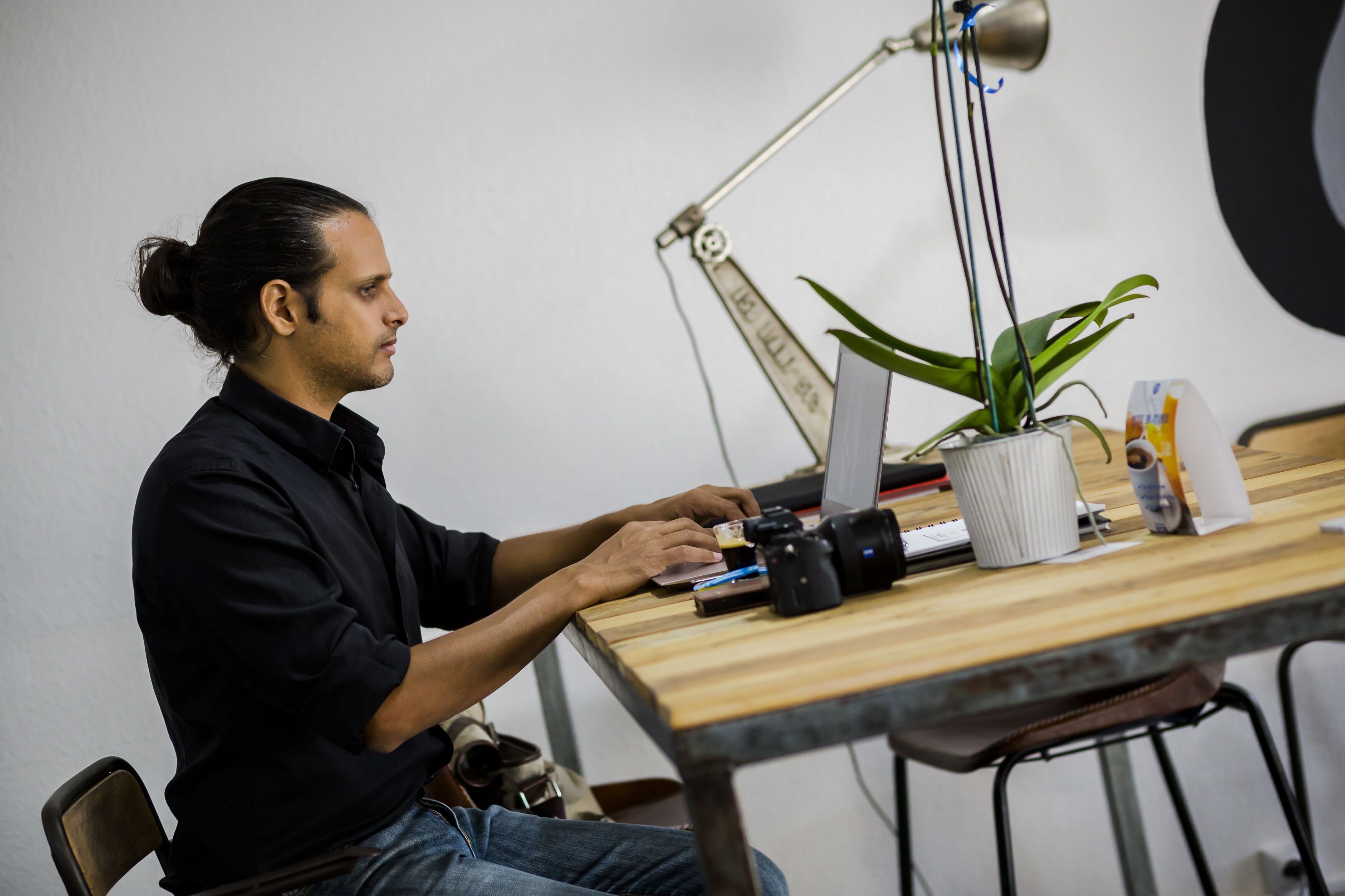 A person seated at a wooden table with a laptop, camera, and plant.