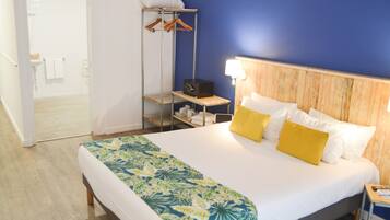A hotel room with a bed, a wooden headboard, a bedside table with a lamp, a shelf with a radio, and a clothing rack.