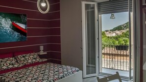 Ein Schlafzimmer mit Bett, einem Gemälde und einem Balkon mit Blick auf eine Stadt und Hügel.