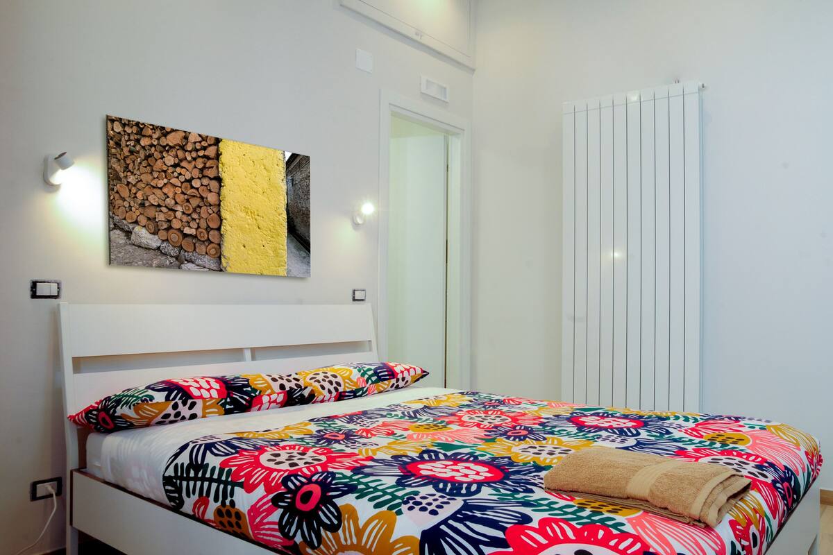 Un lit recouvert d’une couvre-lit à motifs floraux colorés, avec une tête de lit blanche et une œuvre d’art murale représentant des bûches empilées, le tout sur un mur jaune.
