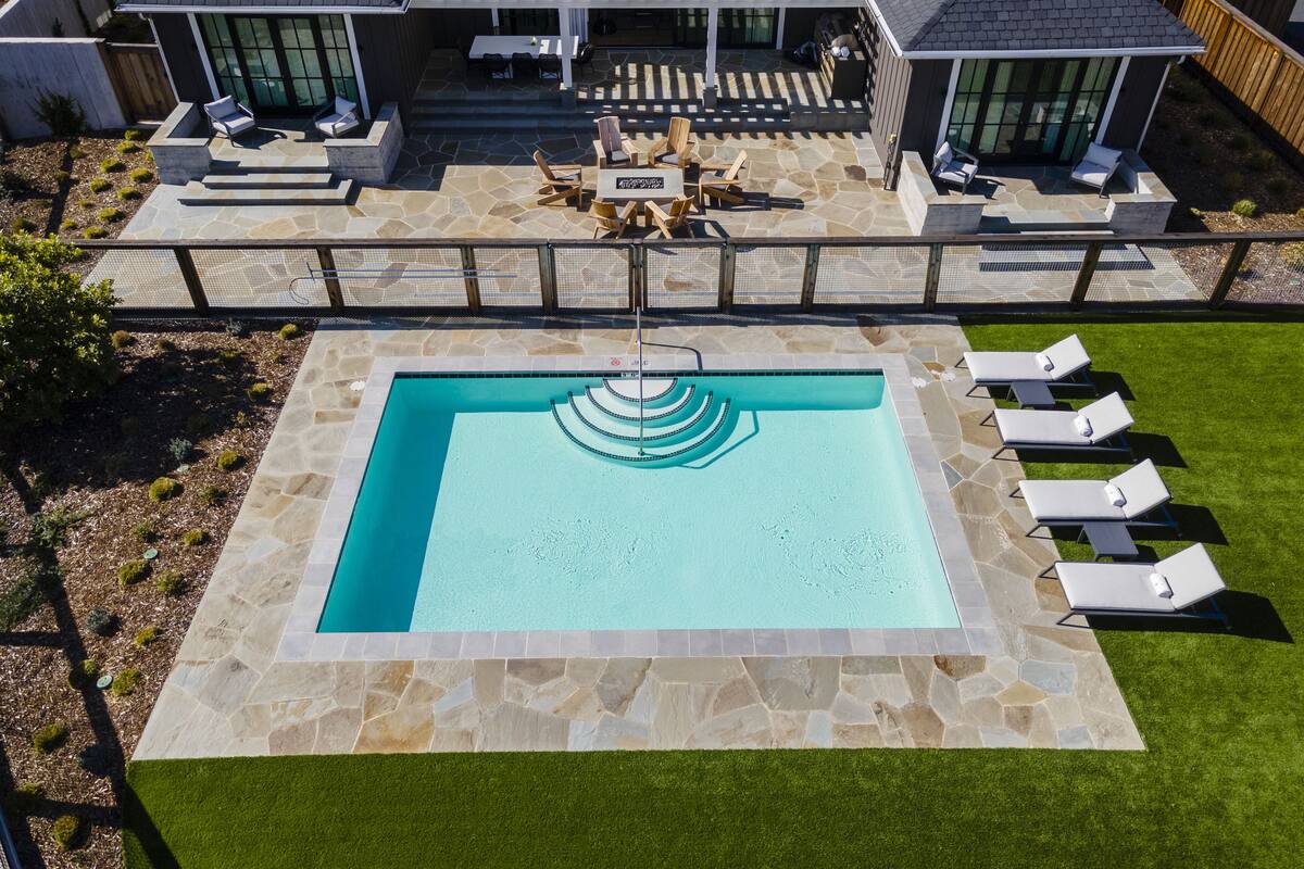 Ein Schwimmbad mit einer Steindeck und Liegestühlen, umgeben von einem gepflegten Rasen und einem Haus mit überdachter Terrasse.