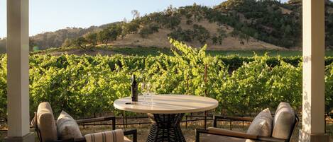 A covered outdoor seating area with a round table and chairs, overlooking a vineyard.